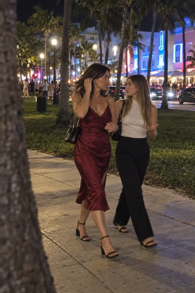 AI-generated mother in a burgundy satin midi dress moving her hair while walking with her adult daughter, in an imagined Ocean Drive night scene.